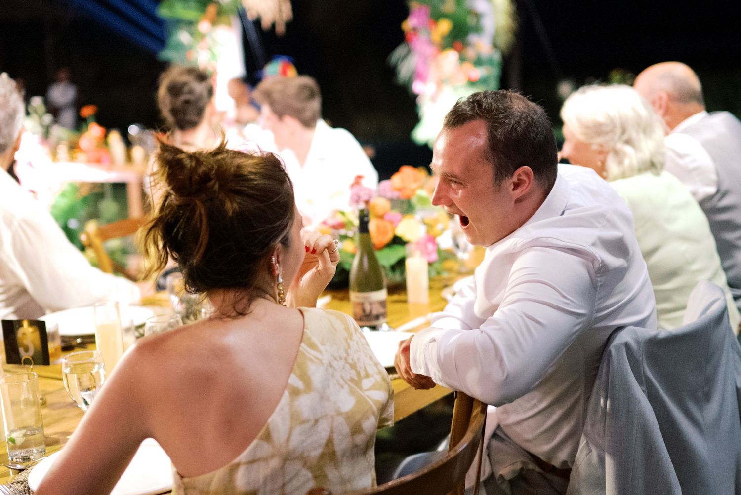 Wedding guests share a joyful laugh at a colorful outdoor tropical reception dinner table at night.