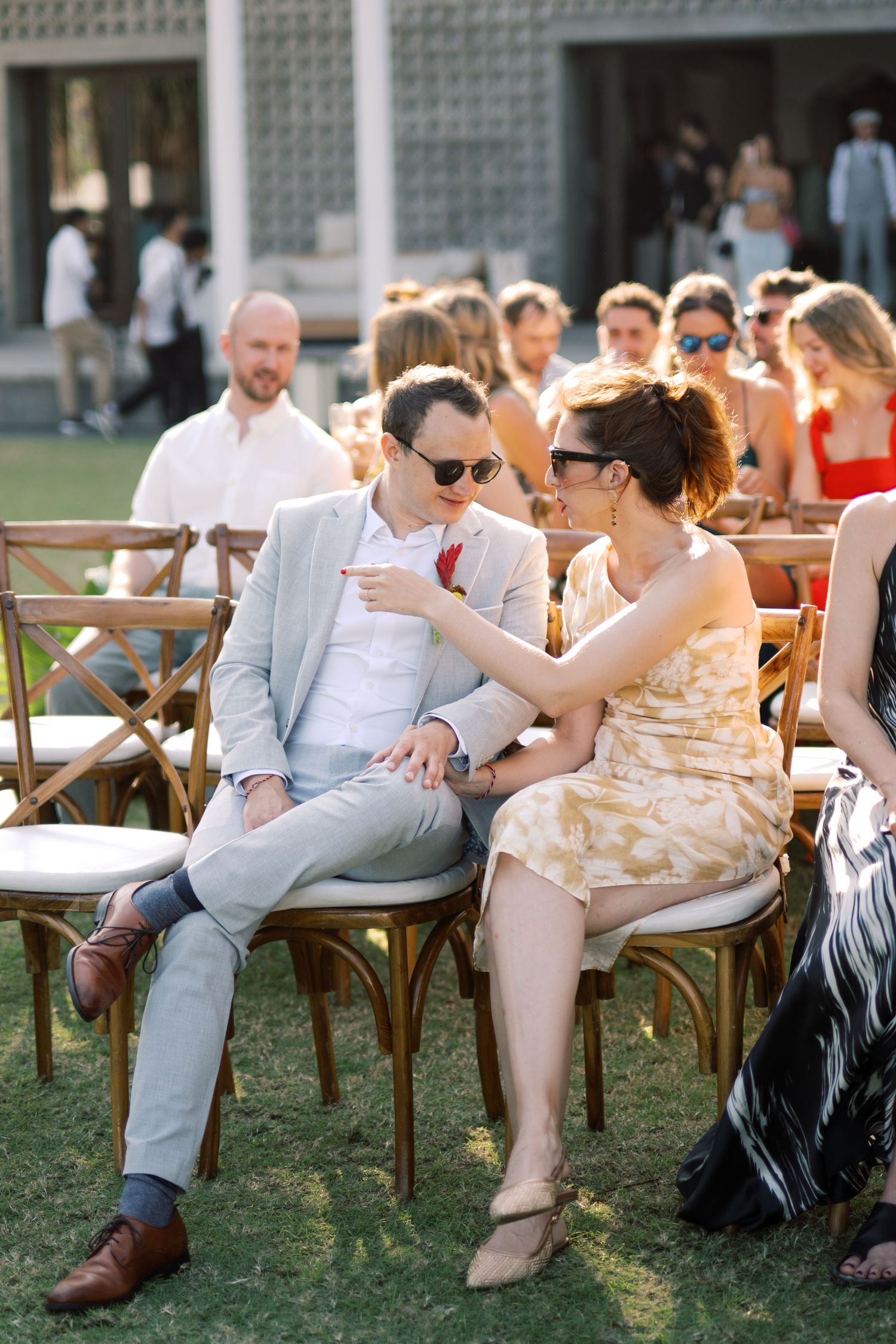 Wedding guests in sunglasses chat and relax on wooden chairs at outdoor ceremony.