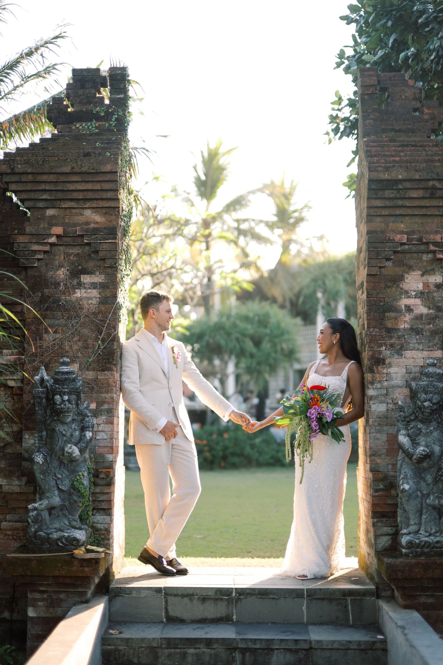 Interracial couple holds hands in a Balinese stone gate surrounded by lush tropical gardens.