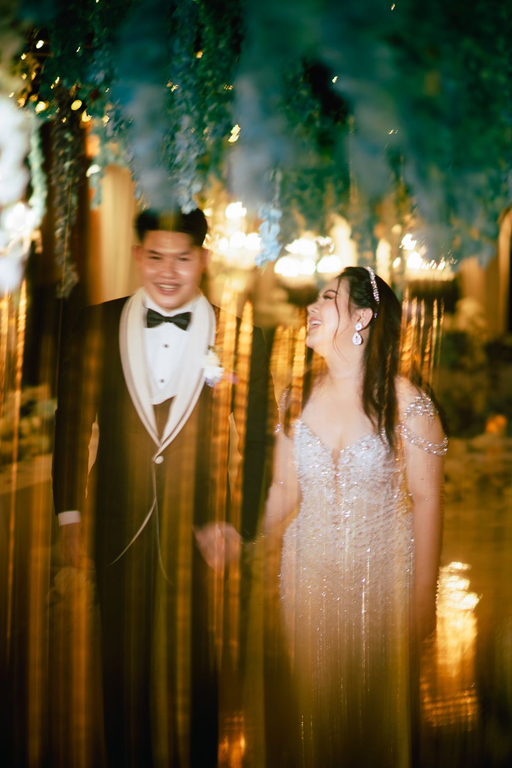 A couple in wedding attire smiles warmly, surrounded by golden bokeh lights and lush greenery in an elegant venue.
