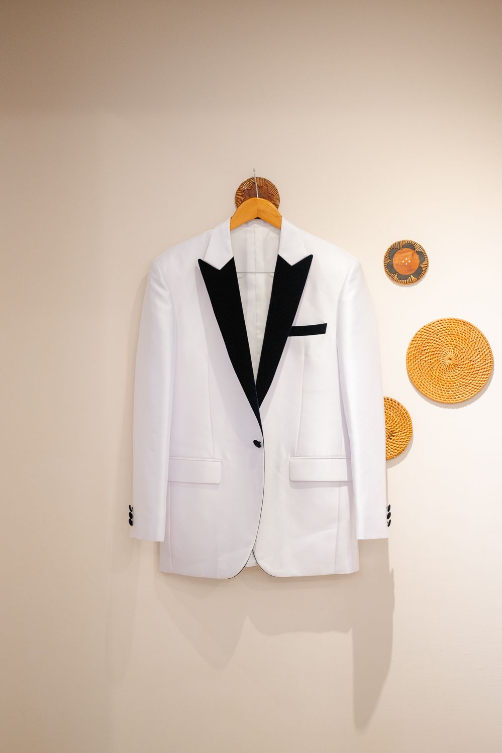 White tuxedo jacket with black lapels hanging on wooden hanger against a neutral beige wall.