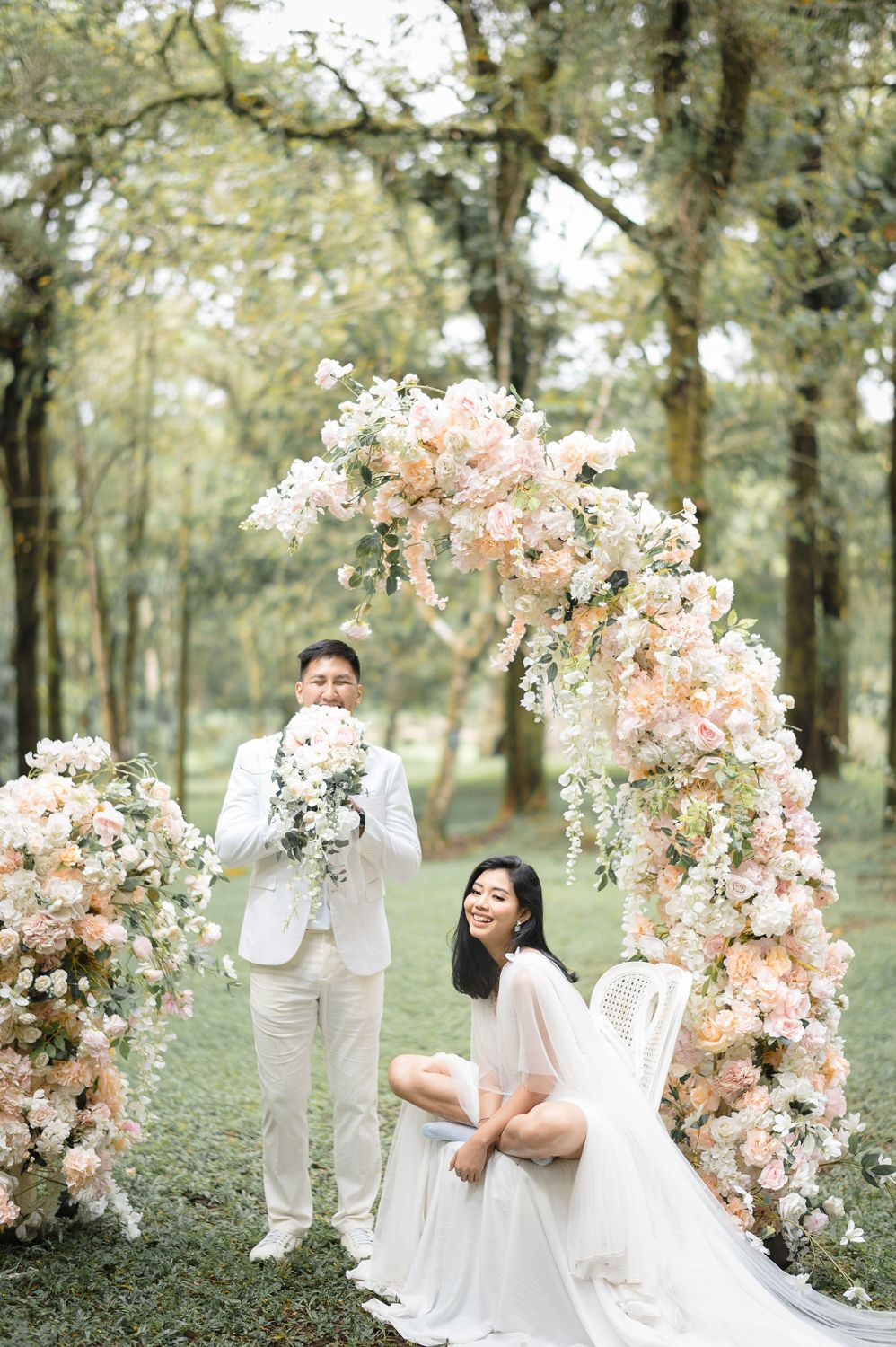 Couple poses under floral arch with peach and white blooms in lush garden wedding setting.