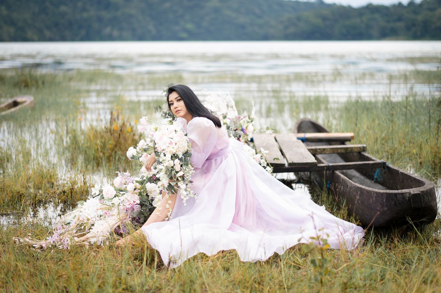 Woman in lavender gown poses with white flowers by a wooden boat near a serene lake surrounded by tall grass.