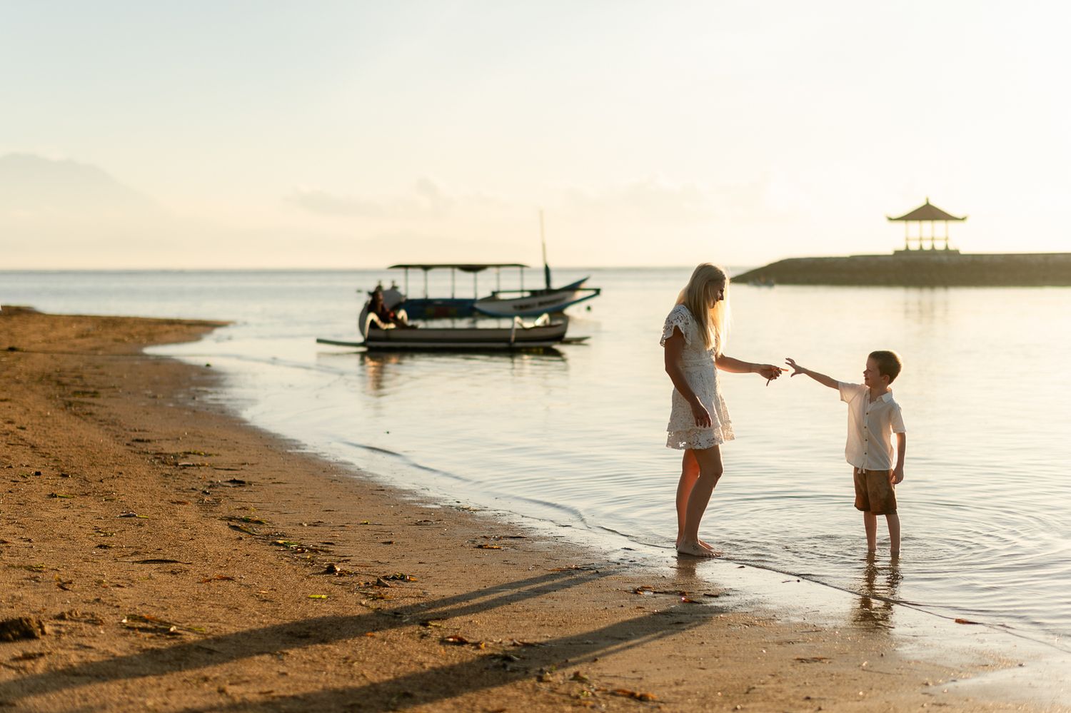 Mother and child holding hands on a tropical beach at sunset with a traditional boat in background.