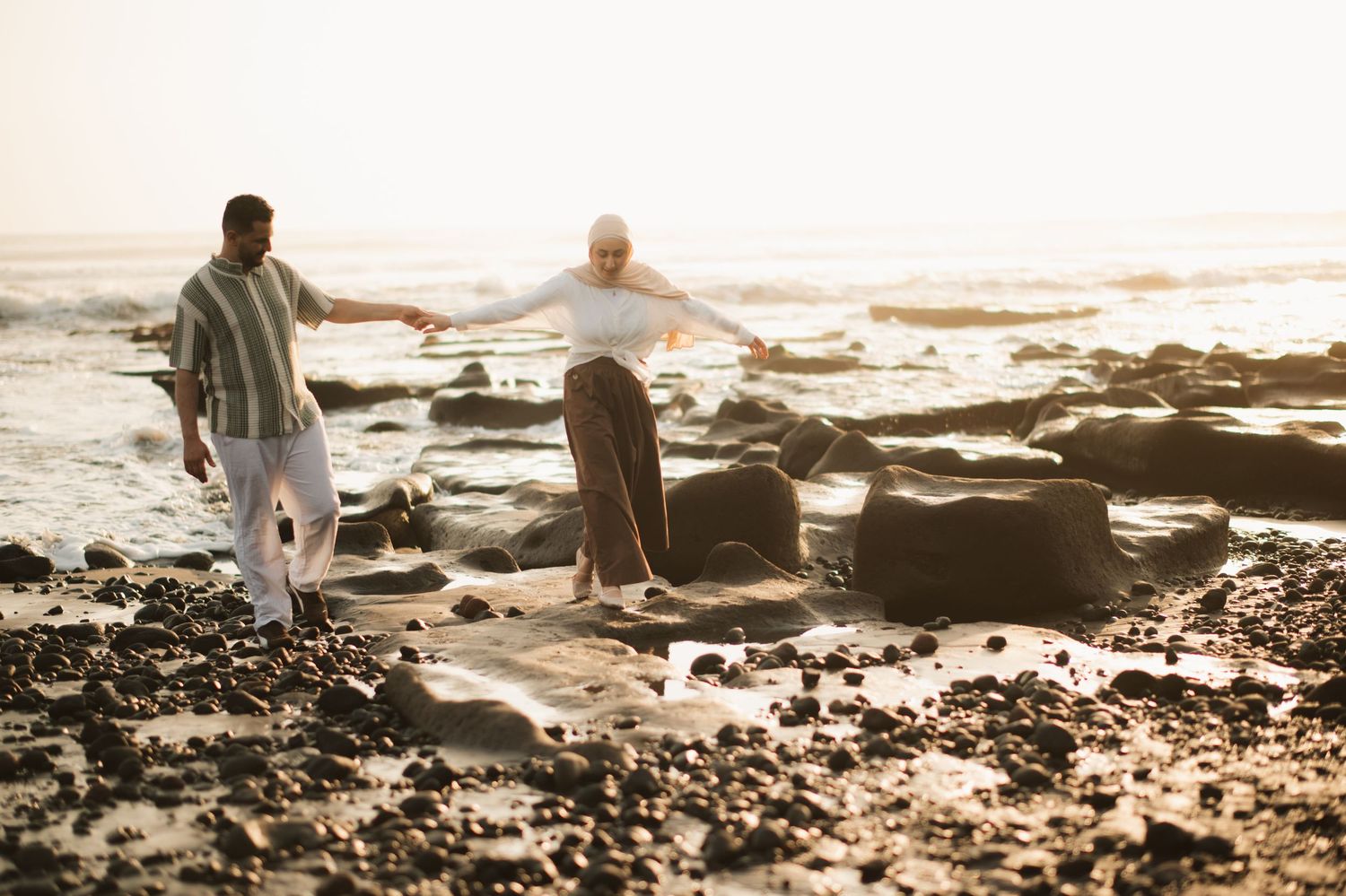 Couple balancing on rocks at a scenic beach during golden hour sunset with ocean waves in the background.