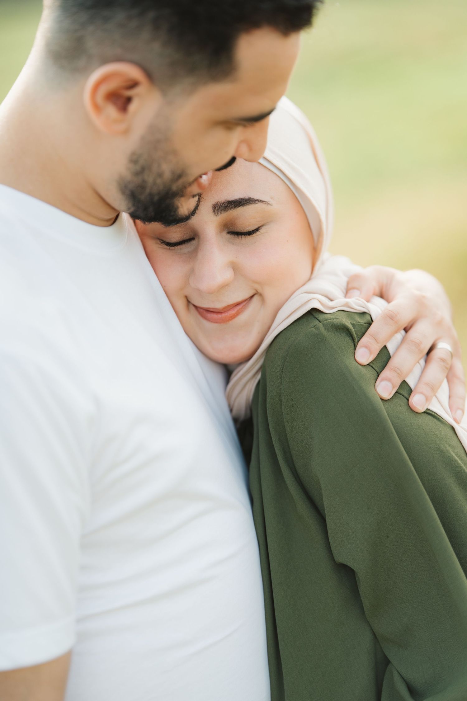 A man in a white t-shirt embraces a smiling woman in a green hijab outdoors in a tender moment.