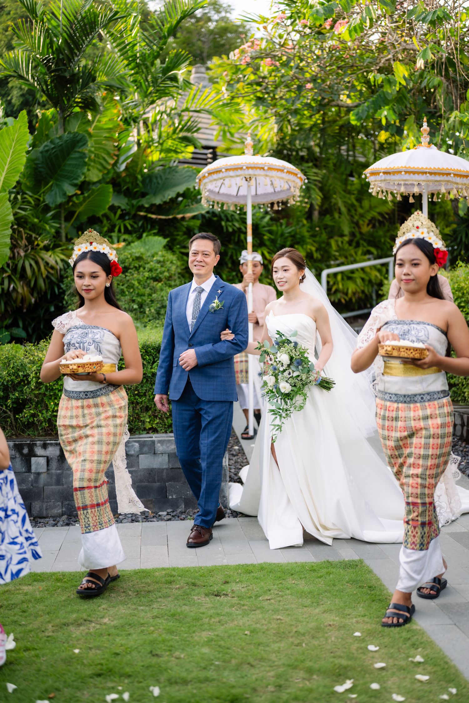 Bride in a white gown walks arm in arm with a man in a blue suit, escorted by attendants in traditional dress with ceremonial umbrellas.