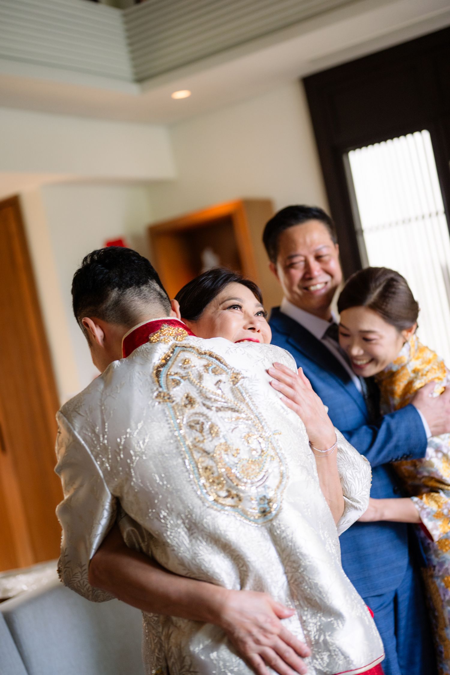 A groom in an embroidered silver Chinese wedding jacket embraces a smiling woman while a man and bride in gold look on joyfully.