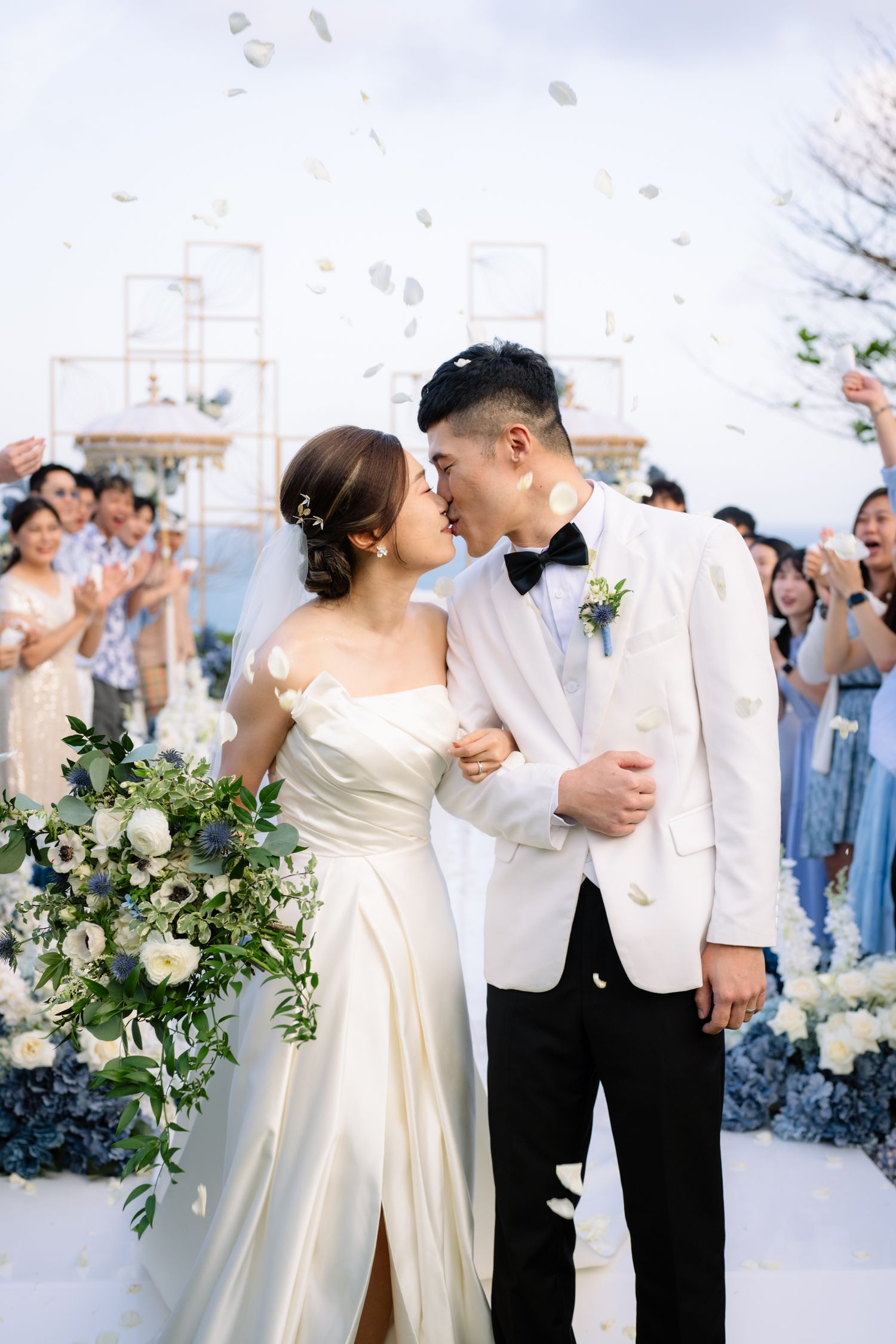 Bride in a satin gown and groom in a white tuxedo kiss during an outdoor wedding ceremony as guests cheer and petals fall.