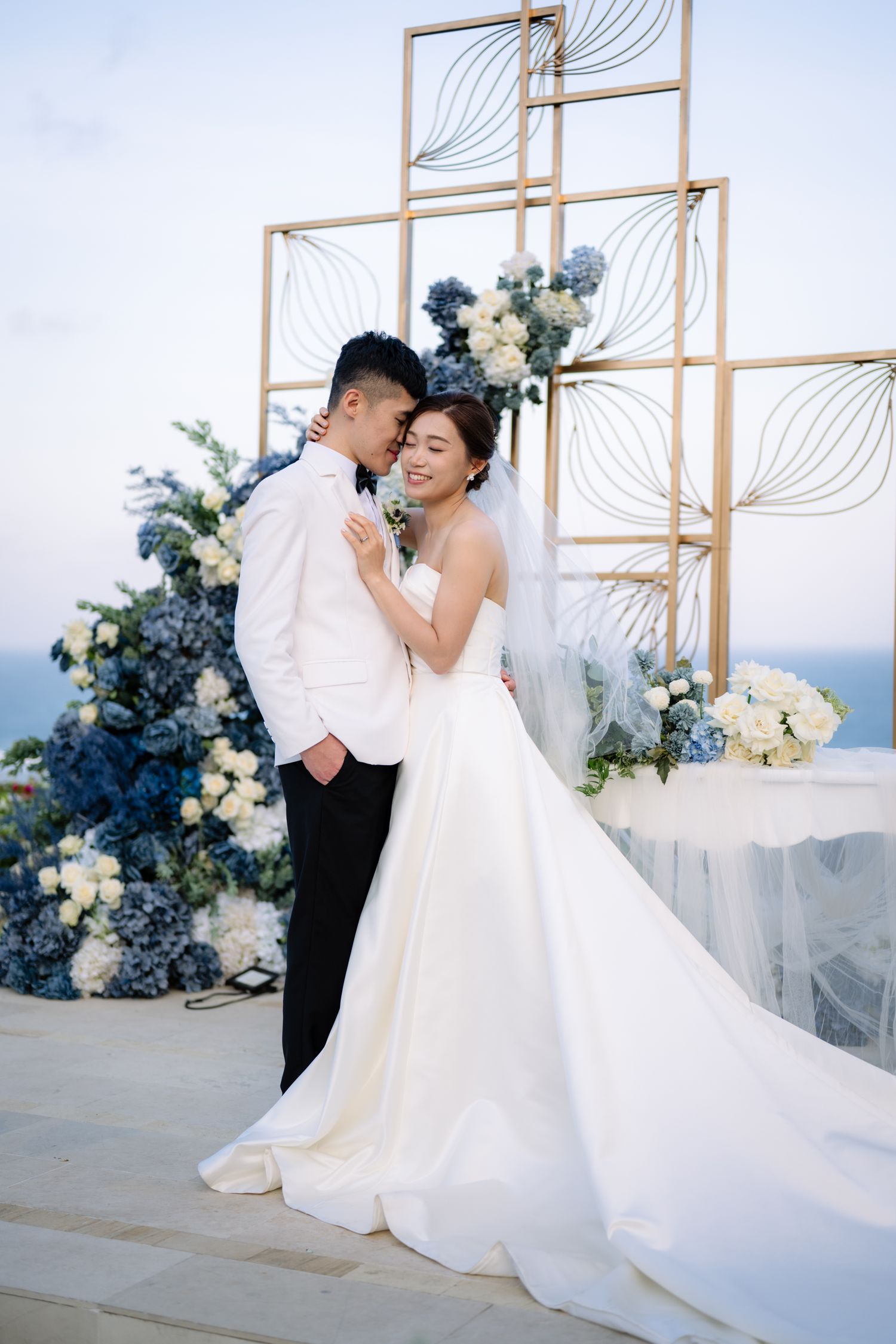 Bride in a white gown and groom in a white tuxedo embrace at a seaside wedding ceremony with blue and white floral arrangements.