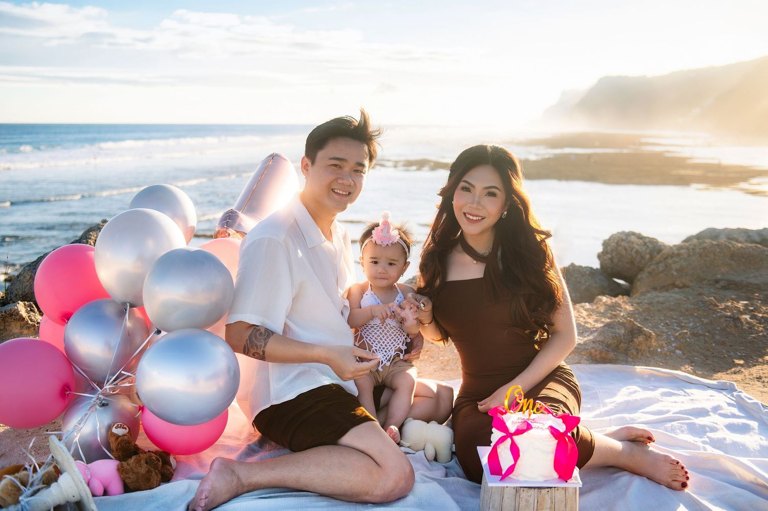 Family celebrating baby’s first birthday on a beach with pink and silver balloons, gifts, and ocean views.