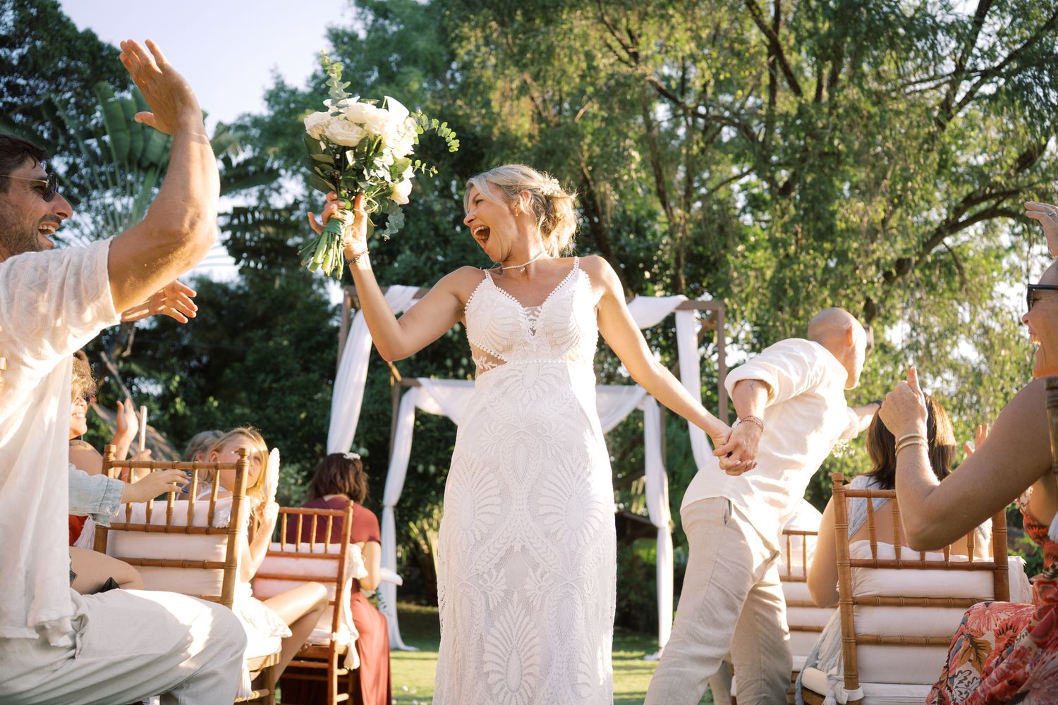 Joyful bride in white lace gown walks down outdoor aisle holding bouquet, guests celebrating around her.