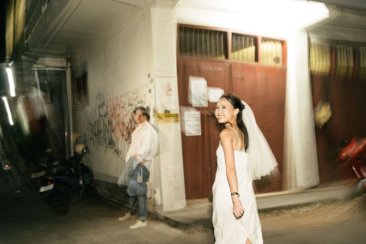A bride in a white dress and veil smiles in an urban alley at night, with a man standing near a red door behind her.