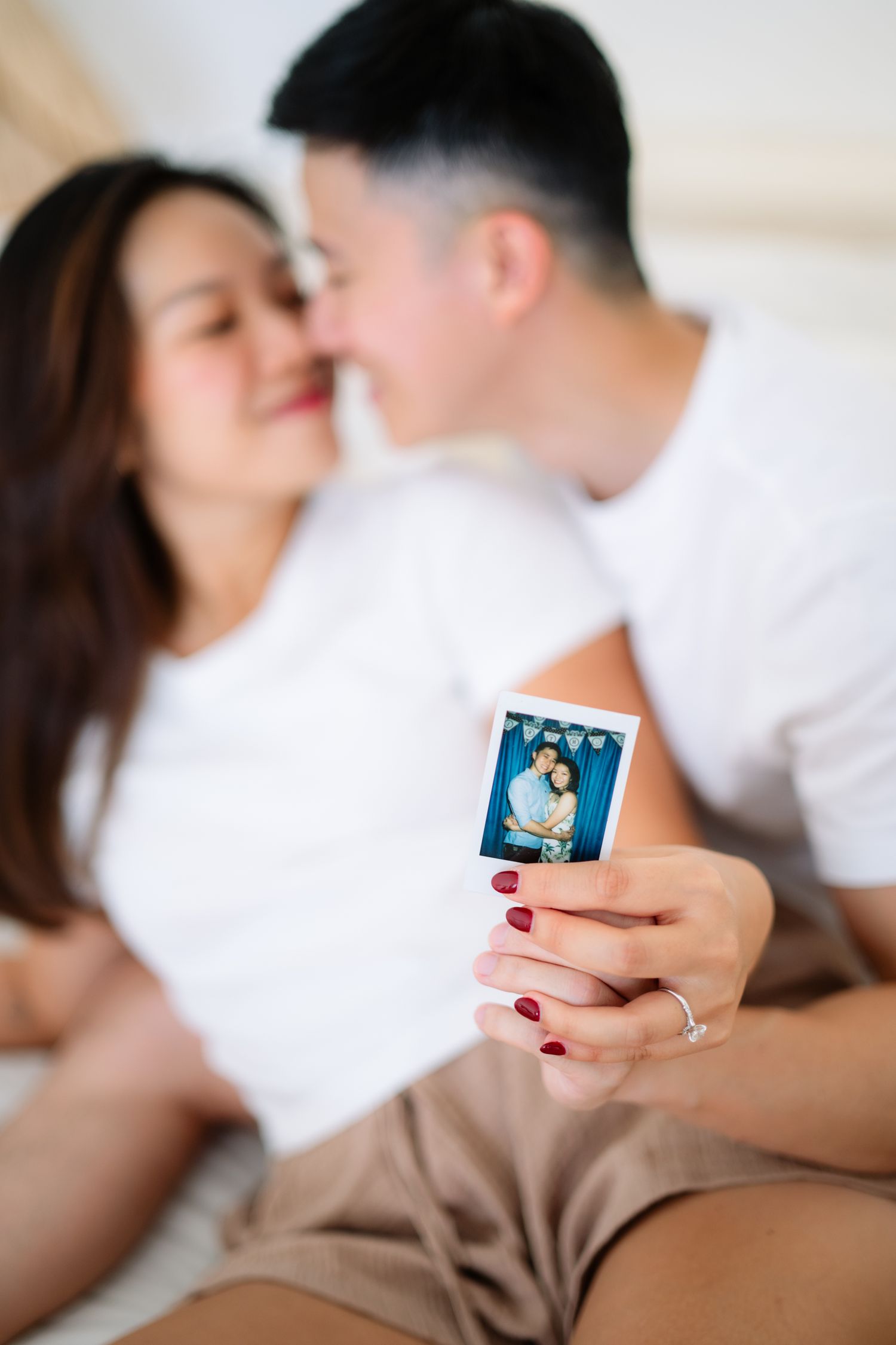 Asian couple sitting on bed, leaning in for a kiss, holding an instant photo of themselves embracing.