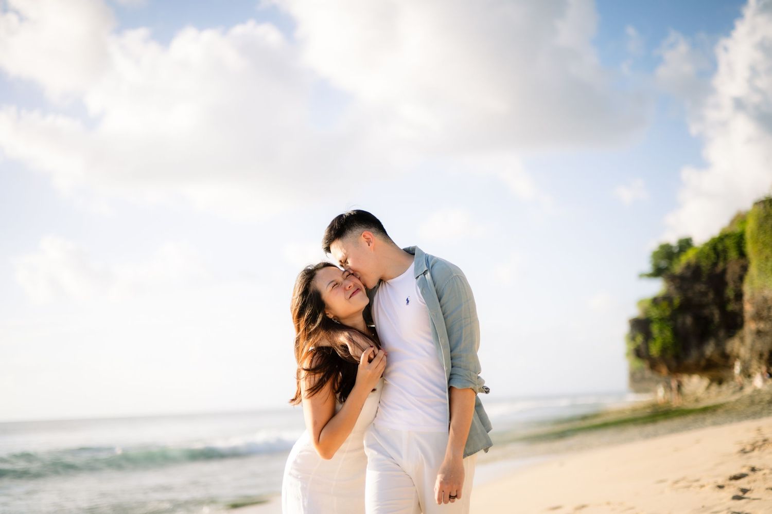 Romantic couple embracing on a tropical beach with turquoise ocean and dramatic cliffs in the background.