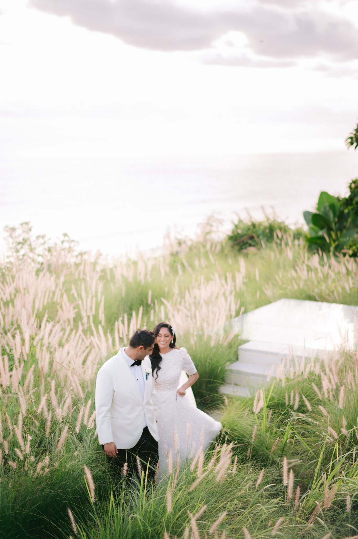 Bride and groom walk through tall golden grass with ocean views at sunset in elegant white wedding attire.