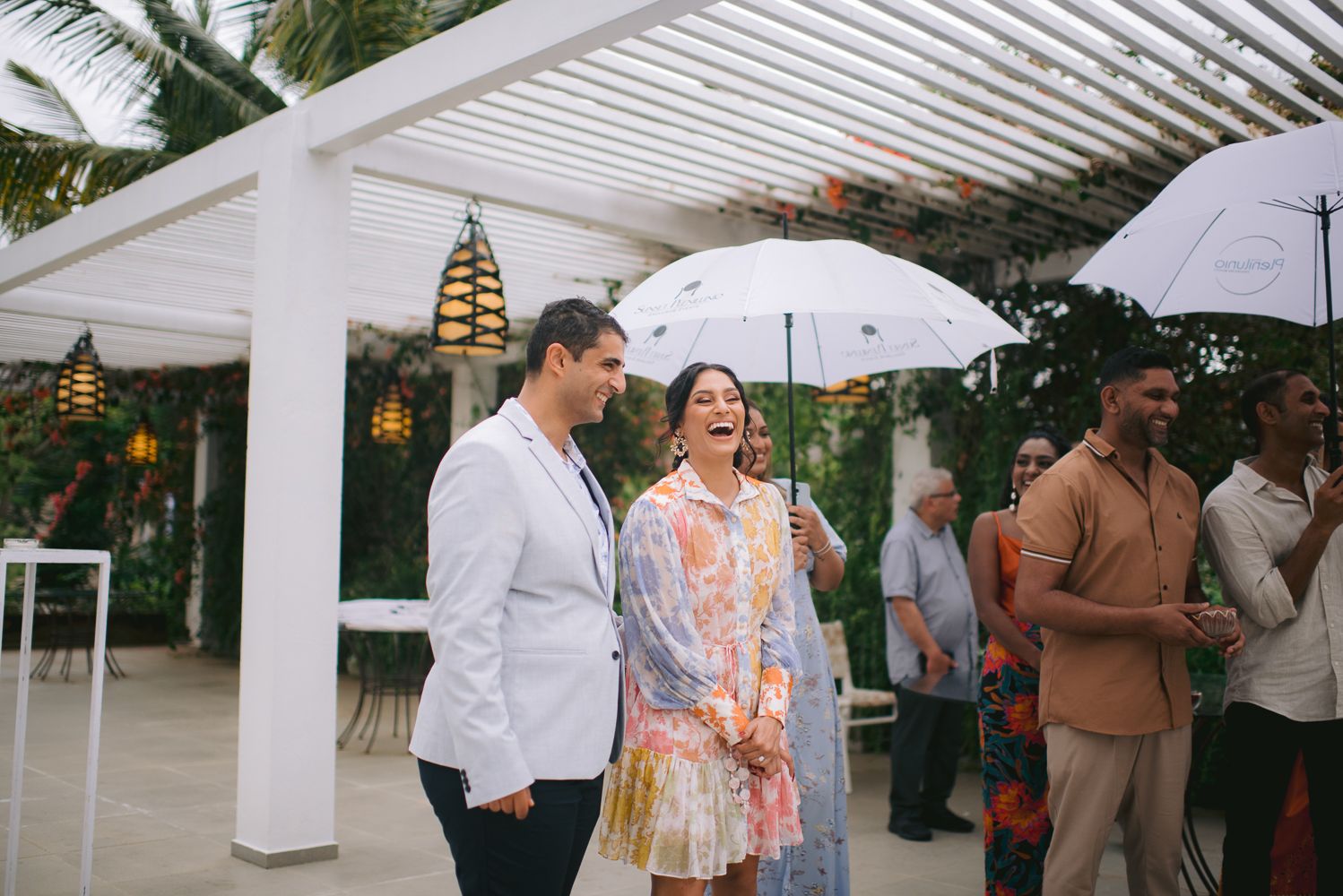 Couple laughing under white umbrella at an outdoor tropical event with guests in colorful attire.