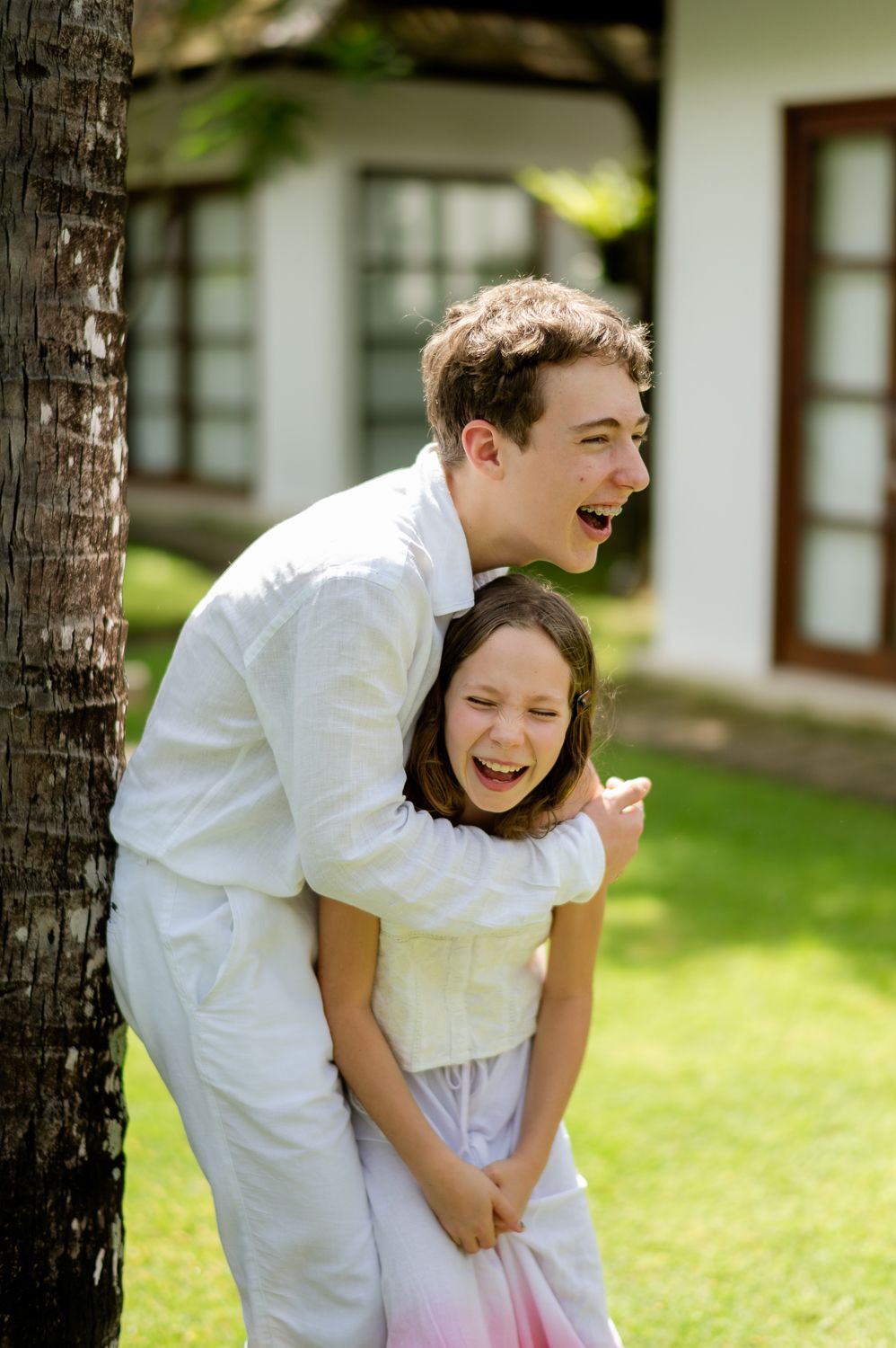 A teenage boy and young girl in white outfits laugh joyfully together outdoors near a tree.