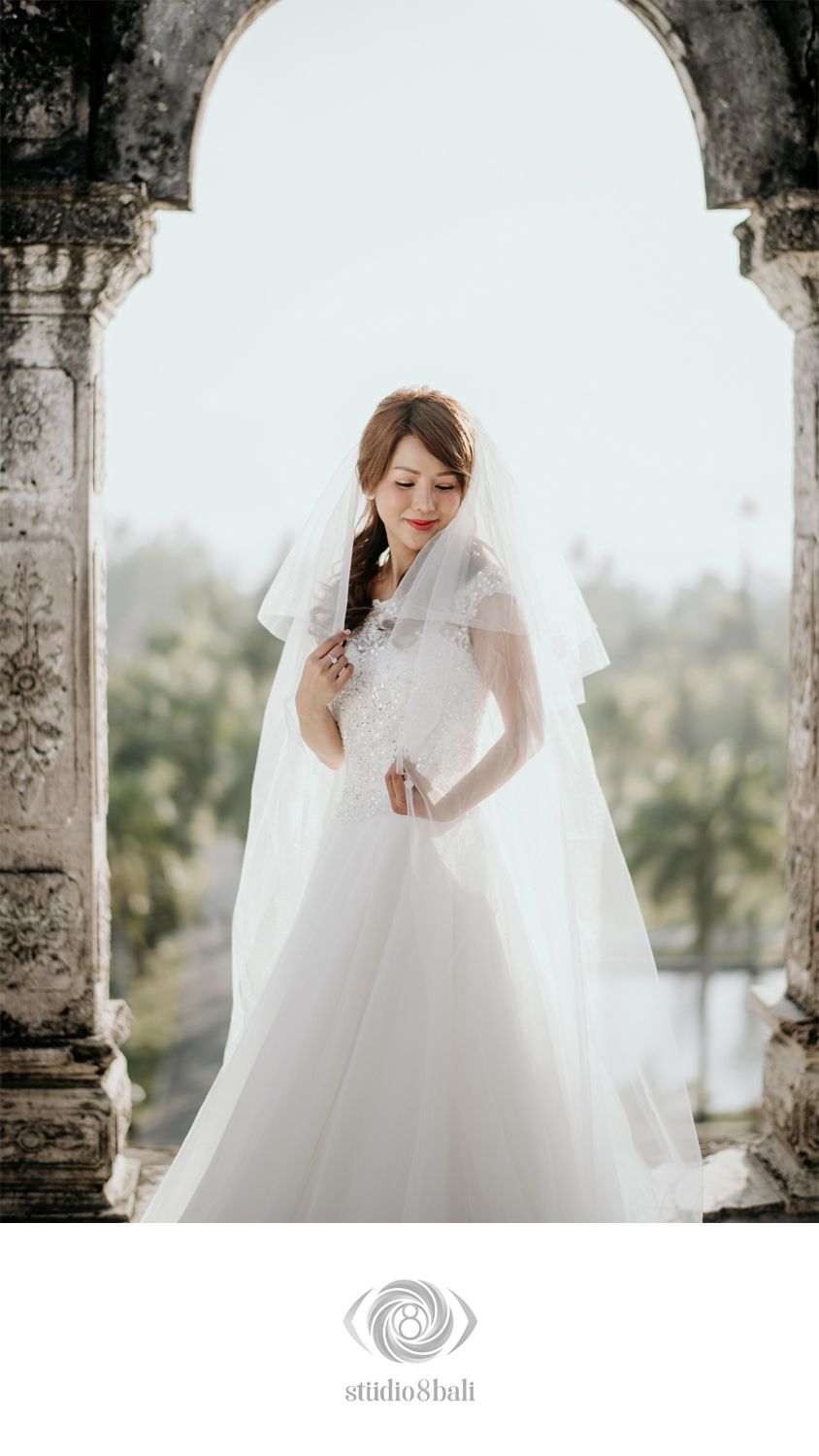Bride in elegant white wedding gown with veil standing in a stone archway with trees in background.