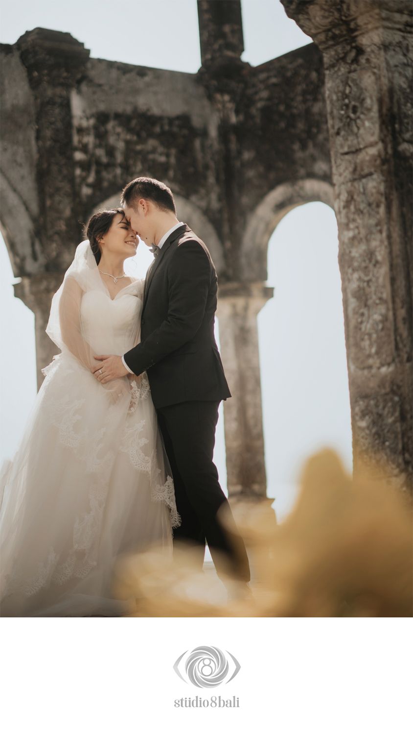 Bride and groom share a romantic moment among ancient stone ruins, bride in white gown, groom in black suit.