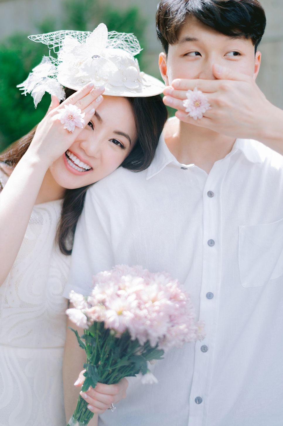 A joyful couple in white attire holds pink flowers up to their faces during a romantic outdoor photo session.