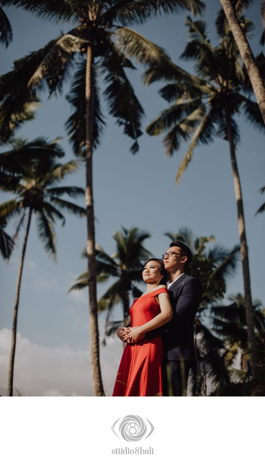 Couple embracing under tall palm trees in Bali, woman in red gown and man in navy suit, romantic pre-wedding photo.