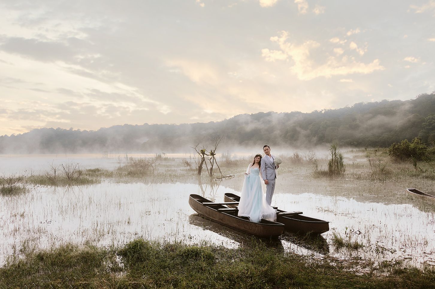 Couple in white attire standing on a wooden boat in a misty lake at dawn.