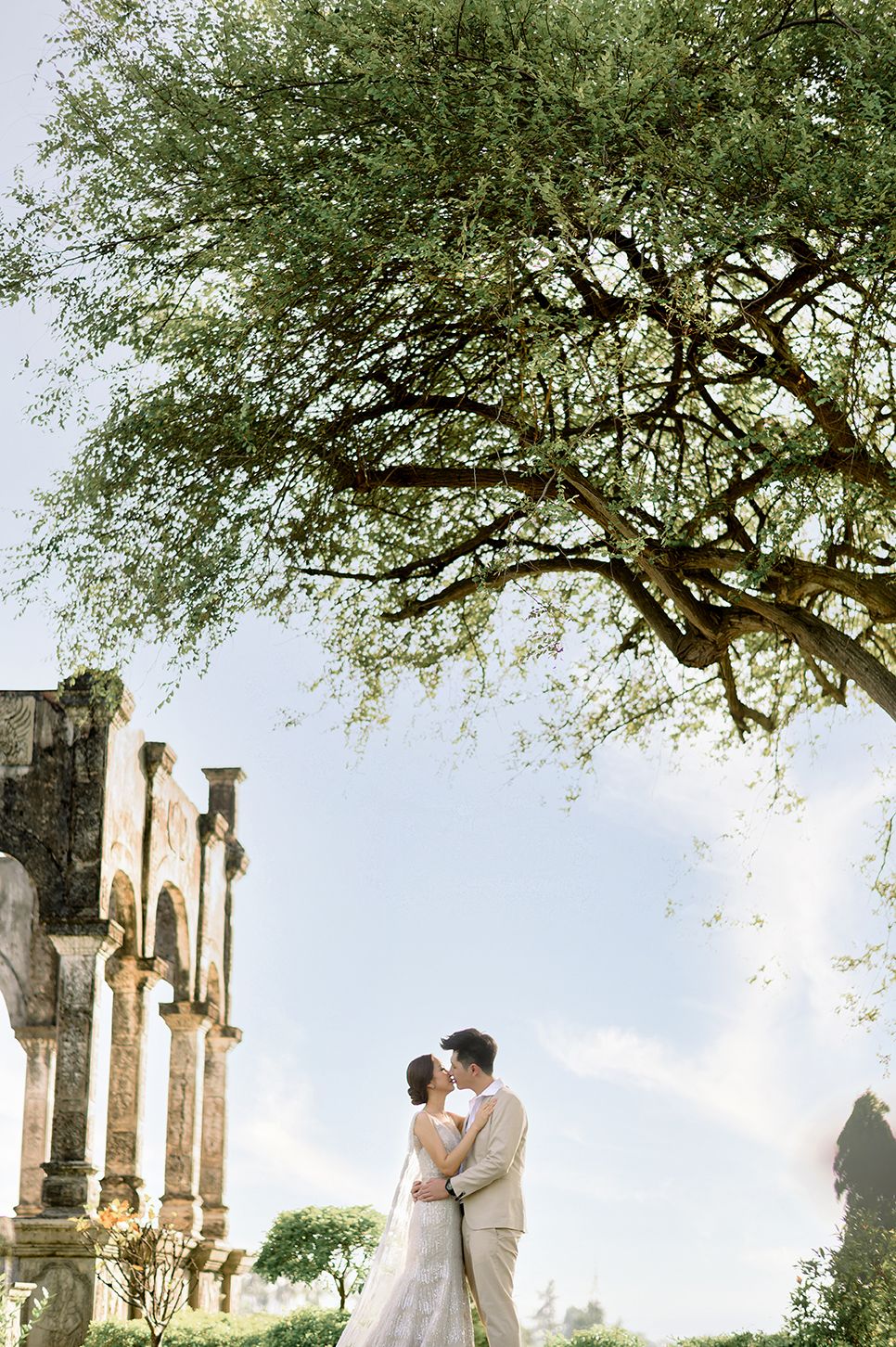 Bride and groom kissing under a large tree beside ancient stone ruins on their wedding day.