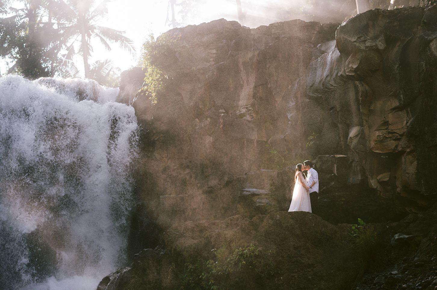 Couple in wedding attire embracing near a stunning tropical waterfall surrounded by misty rocks and lush greenery.