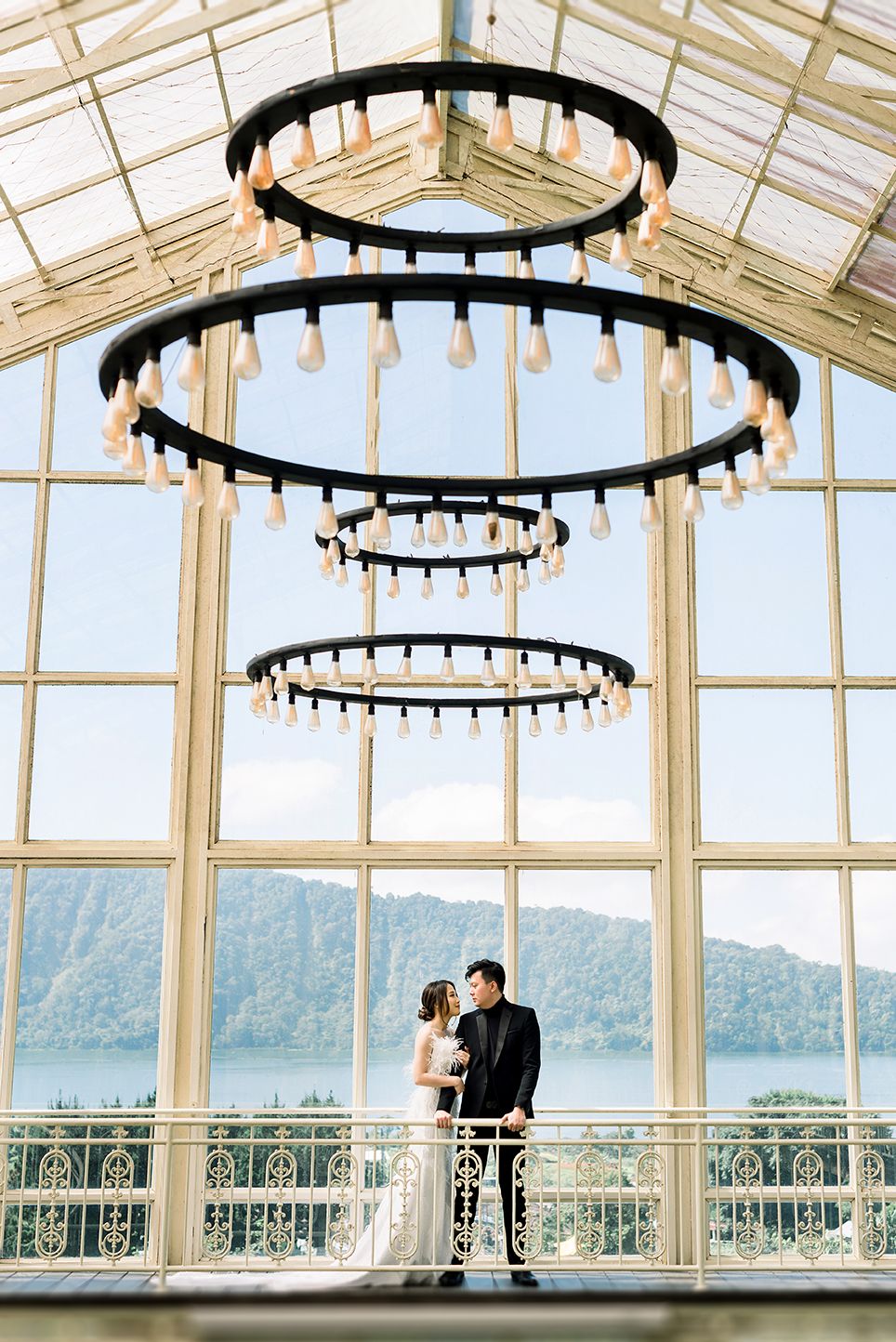 Couple poses under tiered chandelier in glass venue with scenic lake and mountain views on their wedding day.
