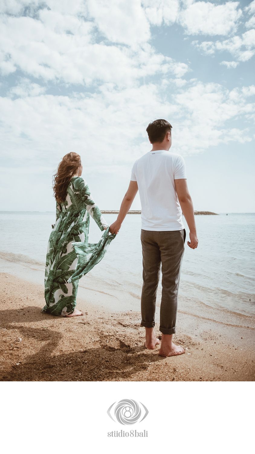 Couple holding hands on a sandy beach shore, woman in floral maxi dress, man in white tee, ocean horizon behind them.