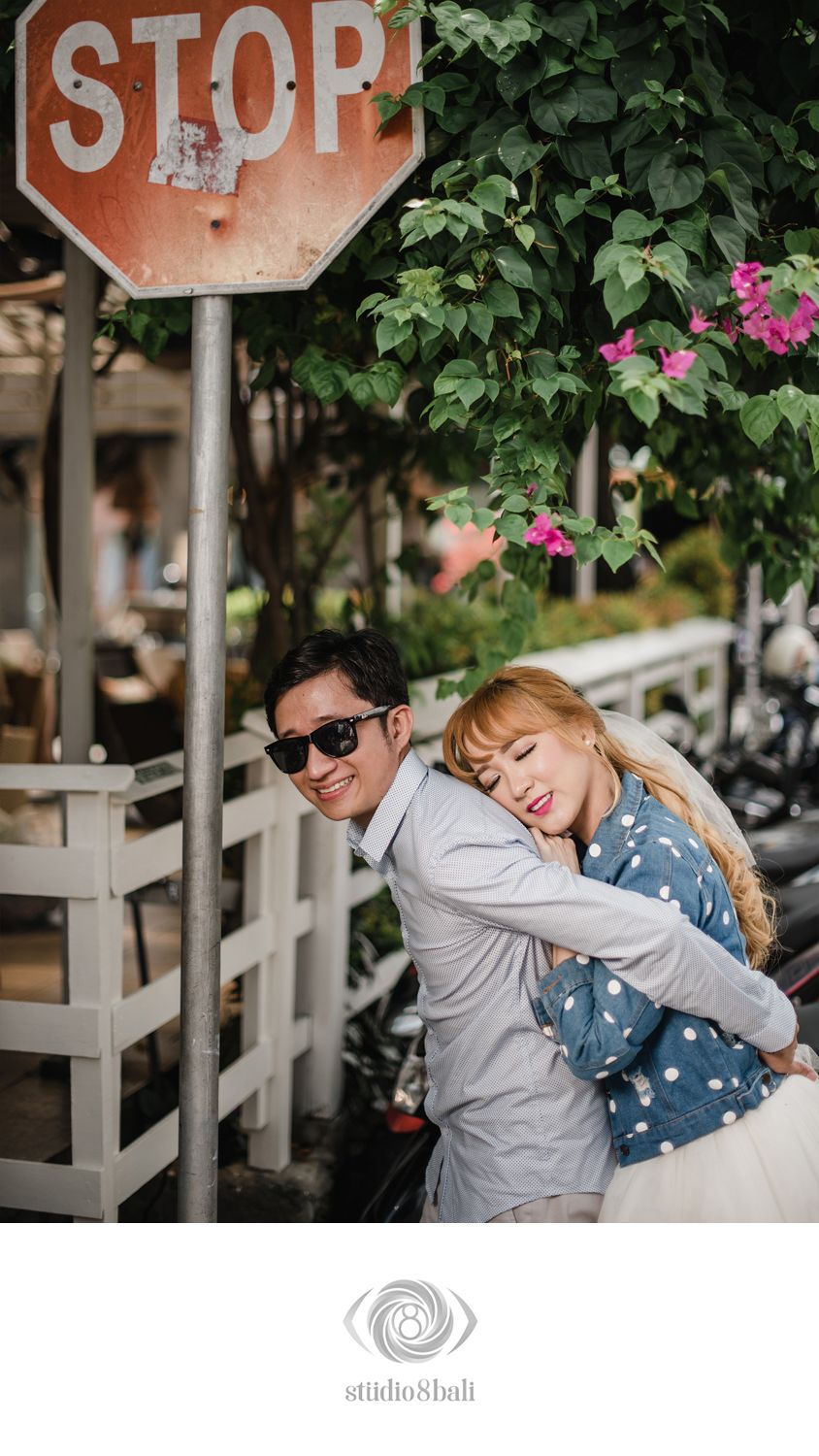 A couple shares a romantic embrace near a vintage stop sign surrounded by lush greenery and pink flowers in Bali.