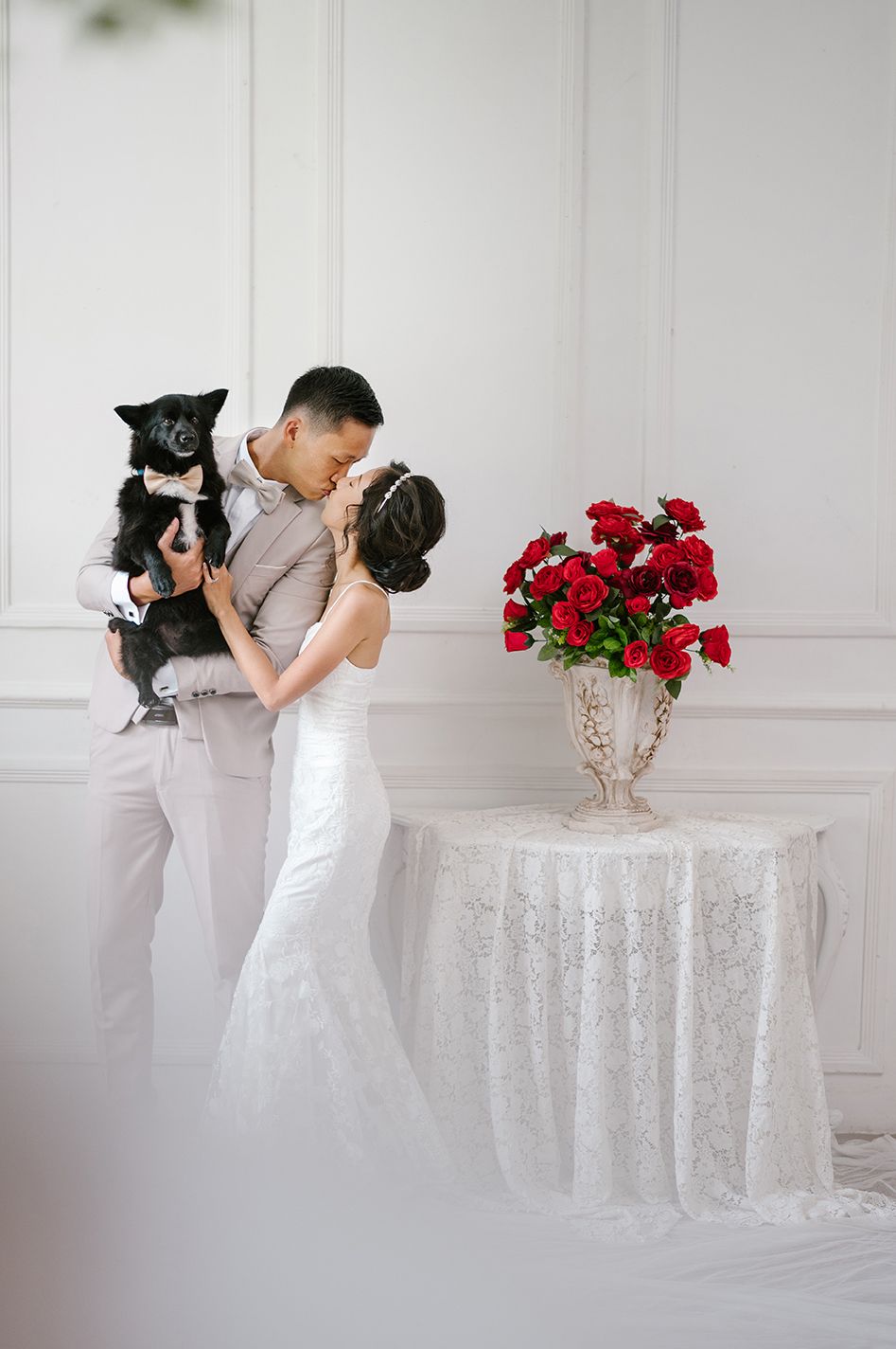 Bride and groom kissing while holding their black dog beside a lace-covered table with red roses in white studio.