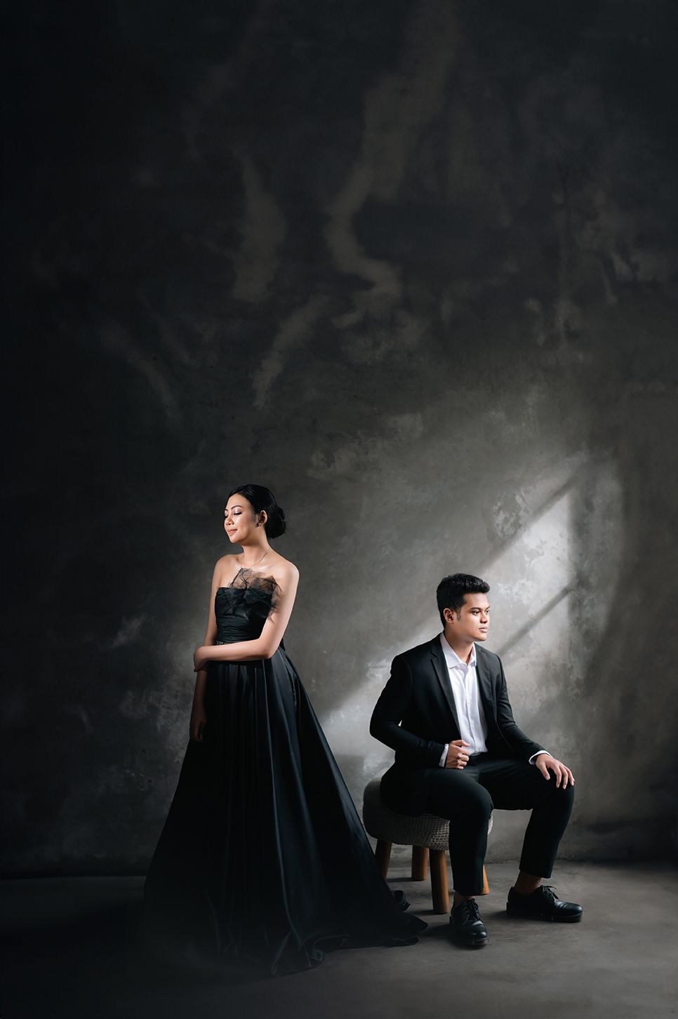 Elegant couple in black formal wear posing dramatically in a moody studio with dramatic lighting and textured gray wall.