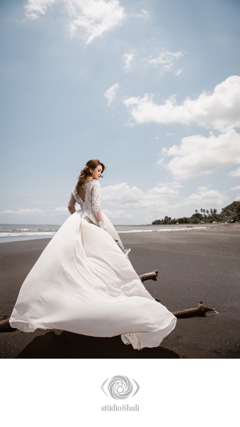 Bride in flowing white lace wedding gown poses on black sand beach with driftwood, dress billowing in ocean breeze.