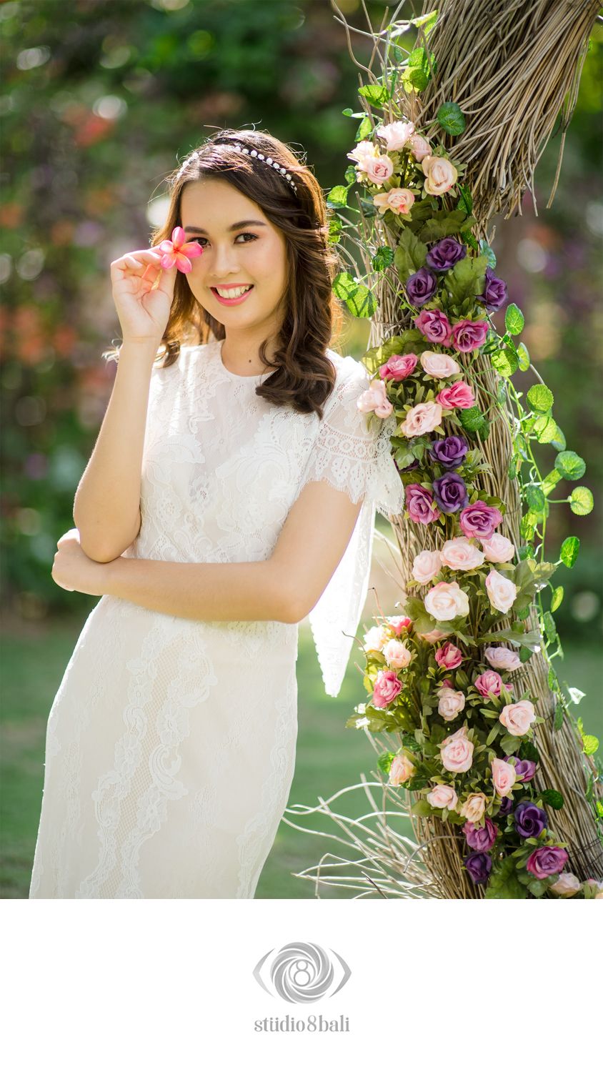 Smiling bride in white lace dress holding pink flower beside floral-decorated palm tree in garden.
