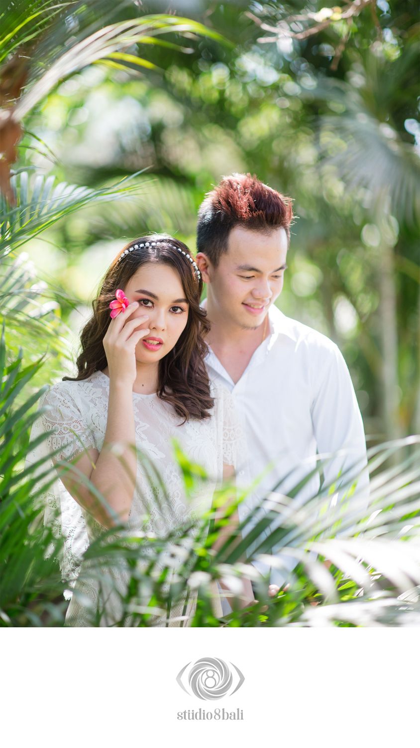 Romantic couple poses among tropical palm leaves, woman with pink flower in hair, both wearing white, by Studio 8 Bali.