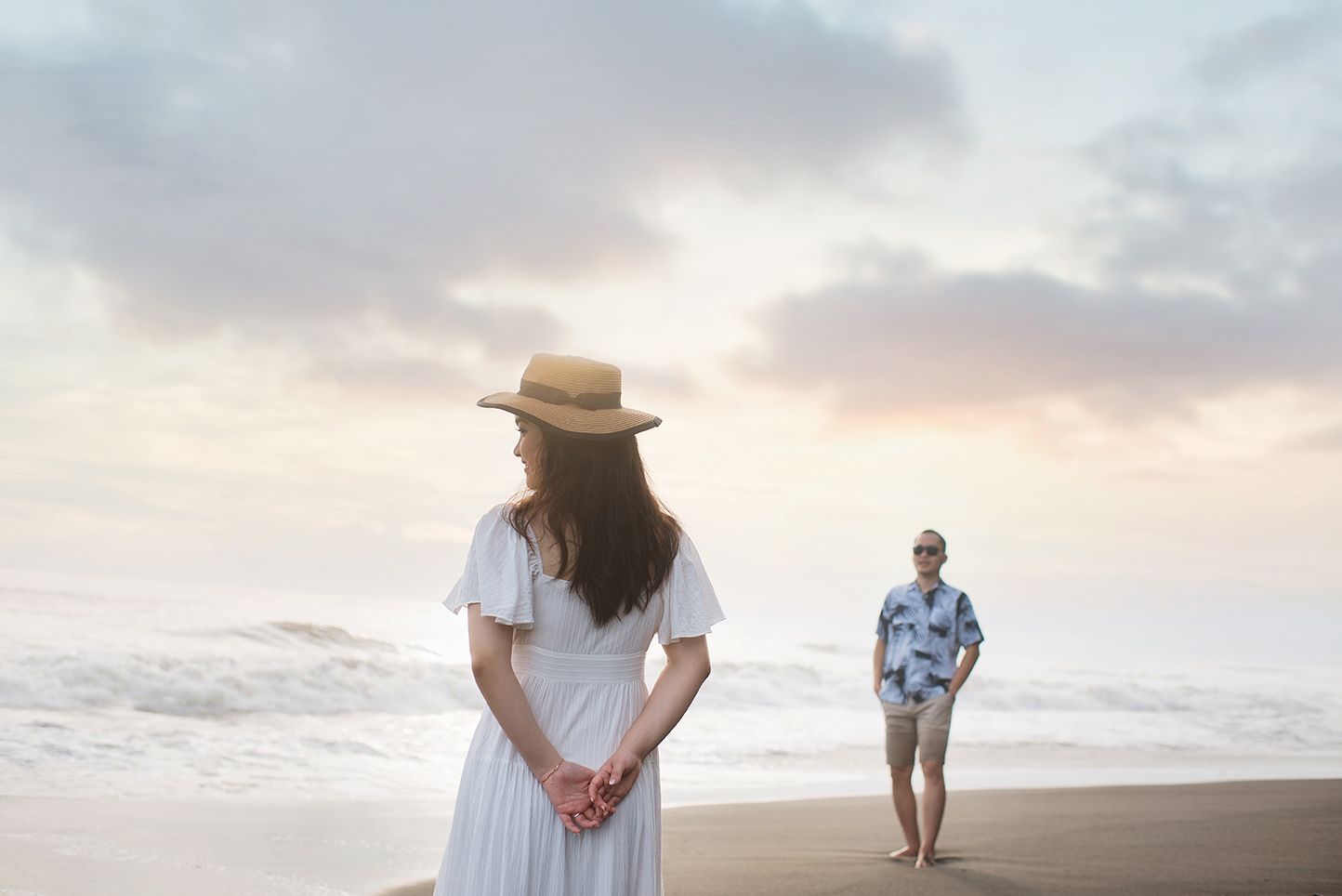 Woman in white dress and hat stands on beach at sunset while a man in Hawaiian shirt walks in background.