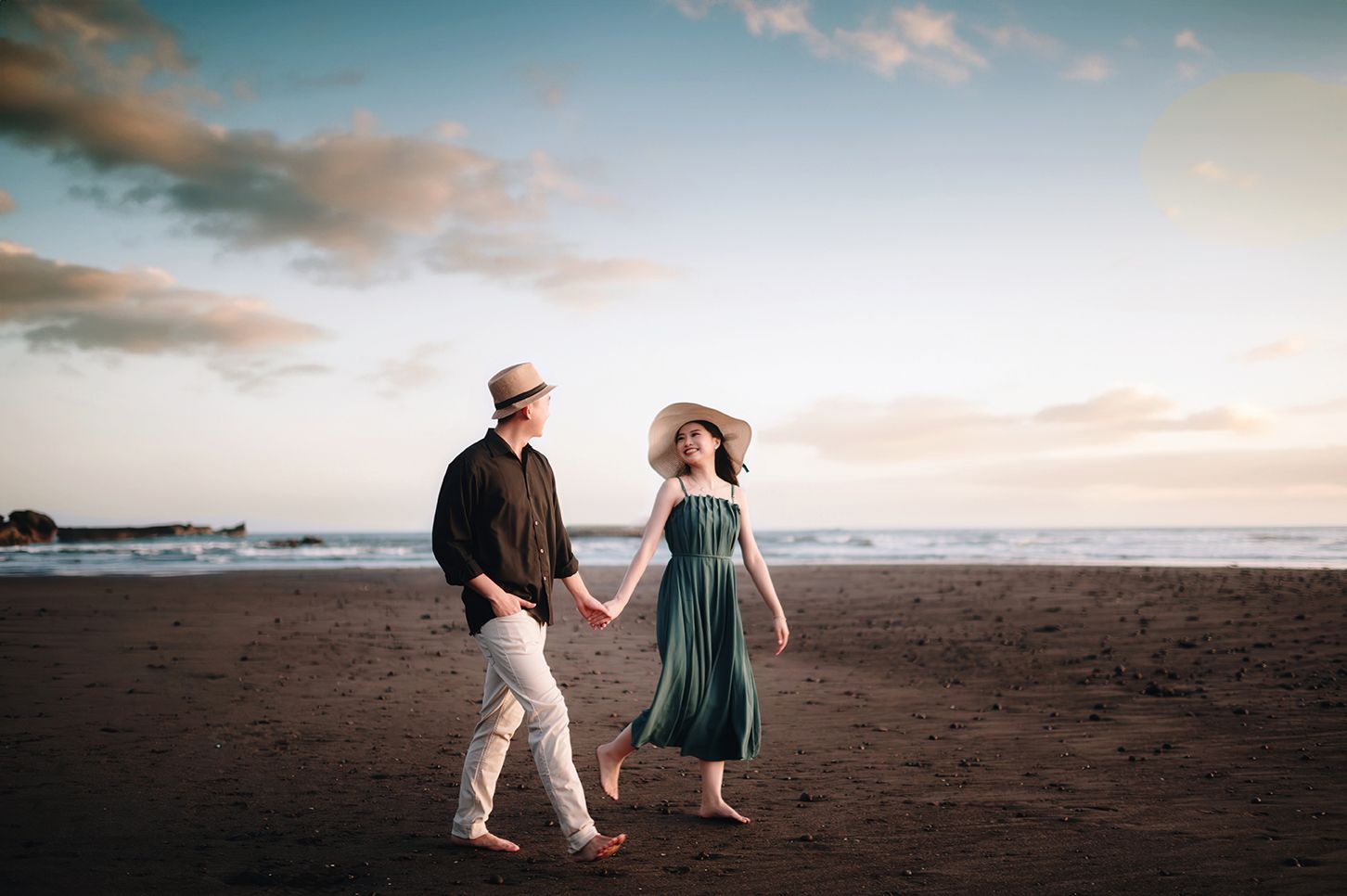 A couple holding hands and walking on a beach at sunset, wearing hats and casual summer attire.