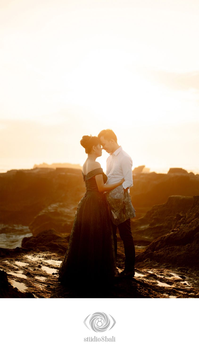 Couple sharing a romantic moment at golden sunset on rocky coastline, woman in black gown, backlit by warm glowing light.