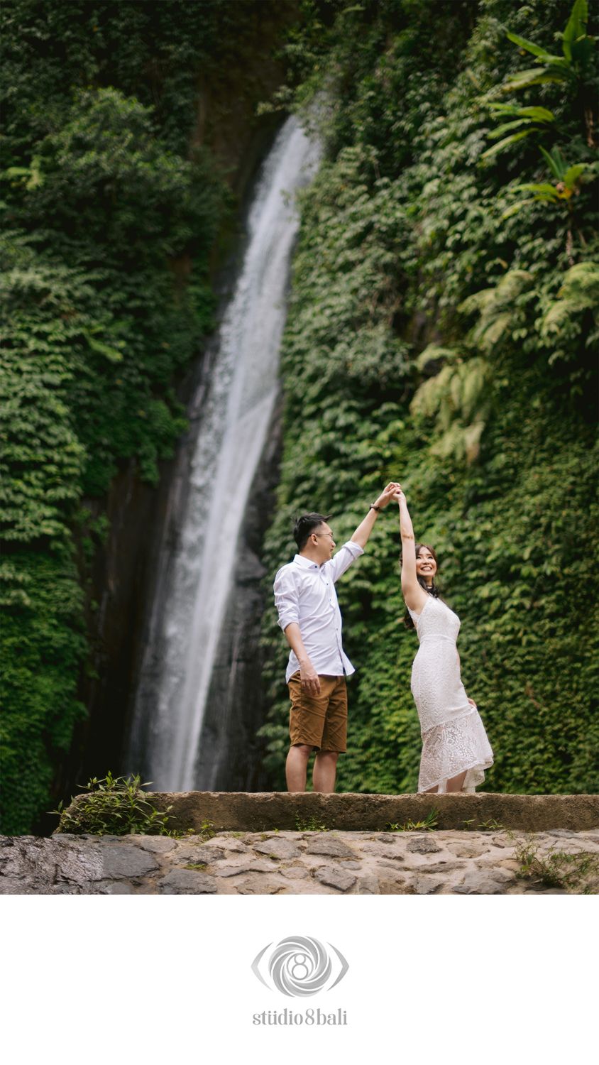 Couple dancing in front of a lush green waterfall in Bali, woman in white dress twirling gracefully.