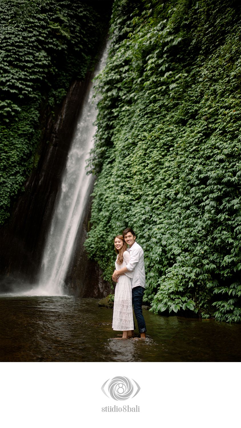 Couple embracing in shallow water before a lush green ivy-covered wall with a cascading waterfall in Bali.