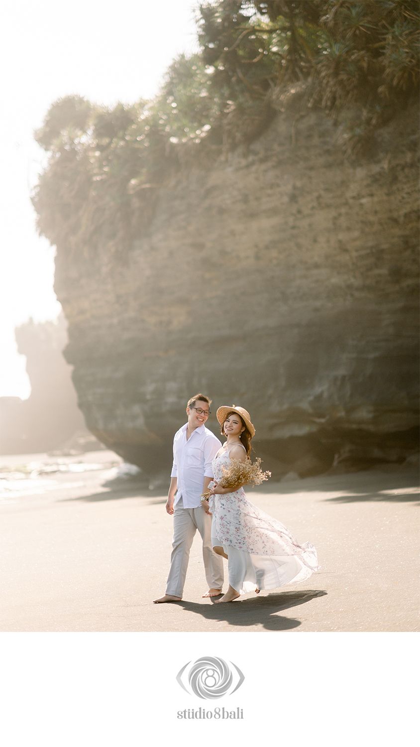 Couple walking on a black sand beach in Bali with dramatic cliffs, soft golden light, floral dress, and bouquet.