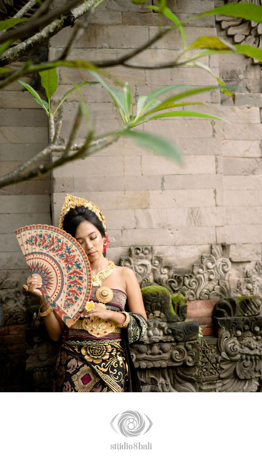 Balinese woman in traditional golden costume holds decorative fan beside ornate stone carvings and tropical plants.