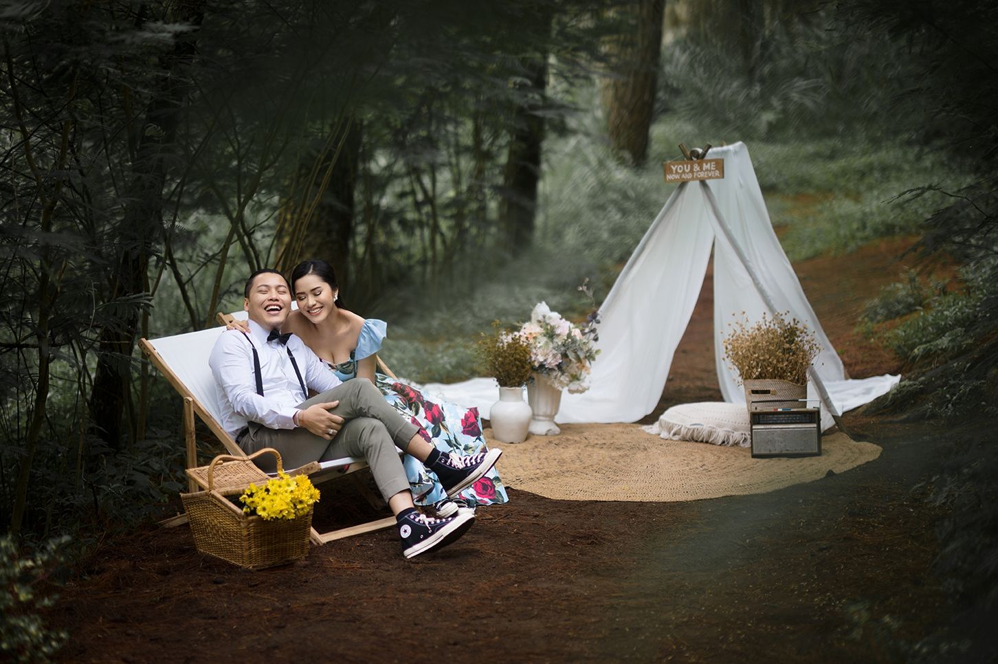 Happy couple relaxing on a lounge chair beside a white teepee tent in a misty forest with yellow flowers and rustic decor.