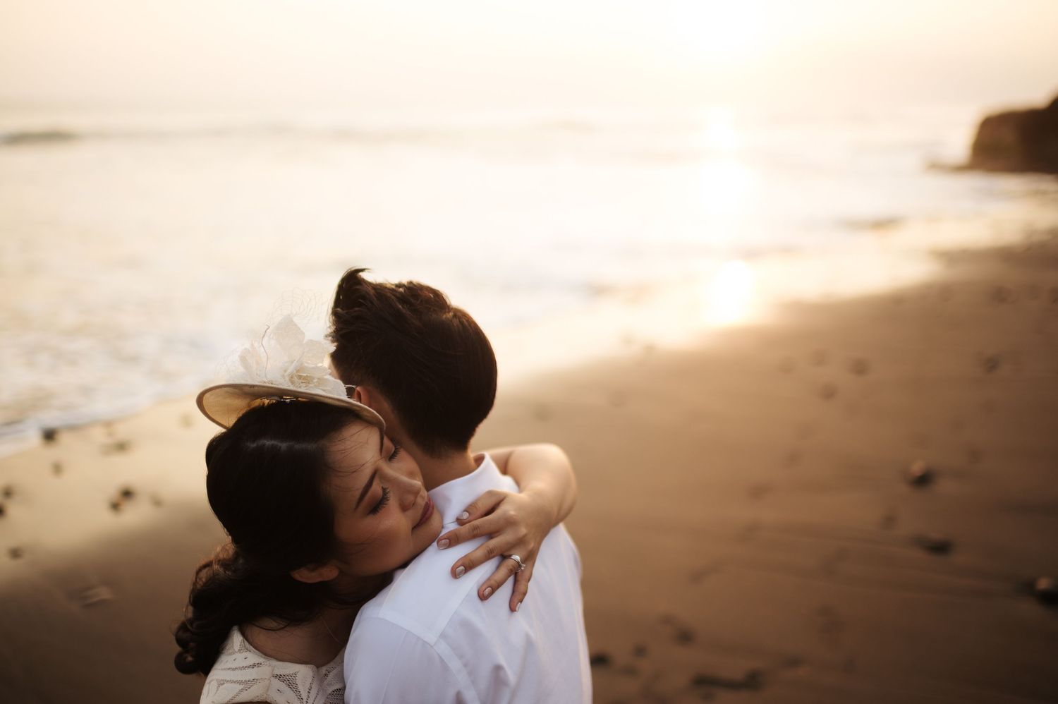 Couple sharing a romantic embrace on a sandy beach at golden sunset, woman wearing a floral hair accessory.
