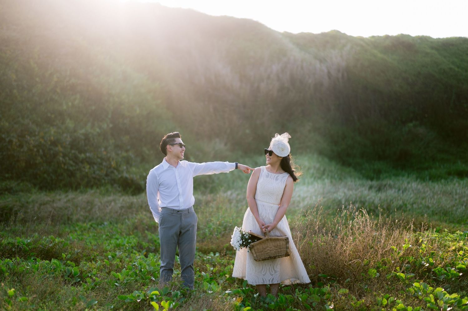 Couple in wedding attire poses in lush green mountain meadow with golden sunlight glowing behind hills.