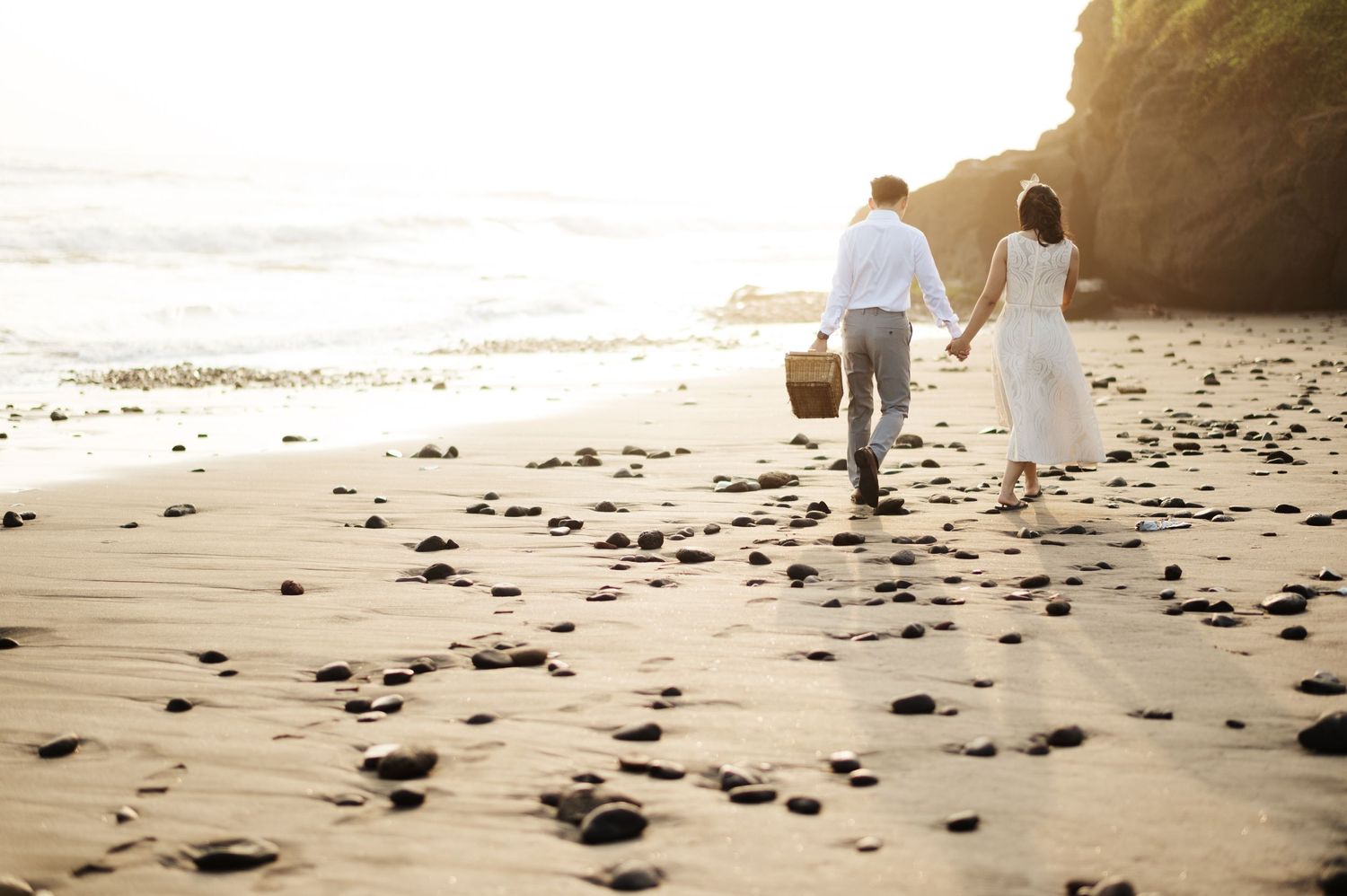 Couple holding hands walking on a rocky beach at sunset, man in white shirt and woman in white dress.