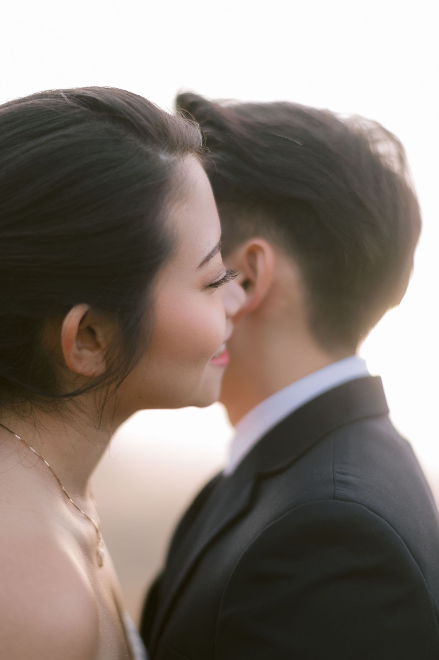 A couple shares a tender kiss in soft golden backlight, the groom in a dark suit and bride with dark upswept hair.