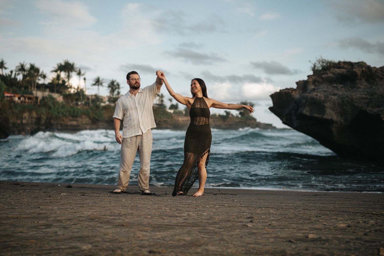 Couple dancing on a tropical beach at sunset with dramatic rock formations and ocean waves in the background.